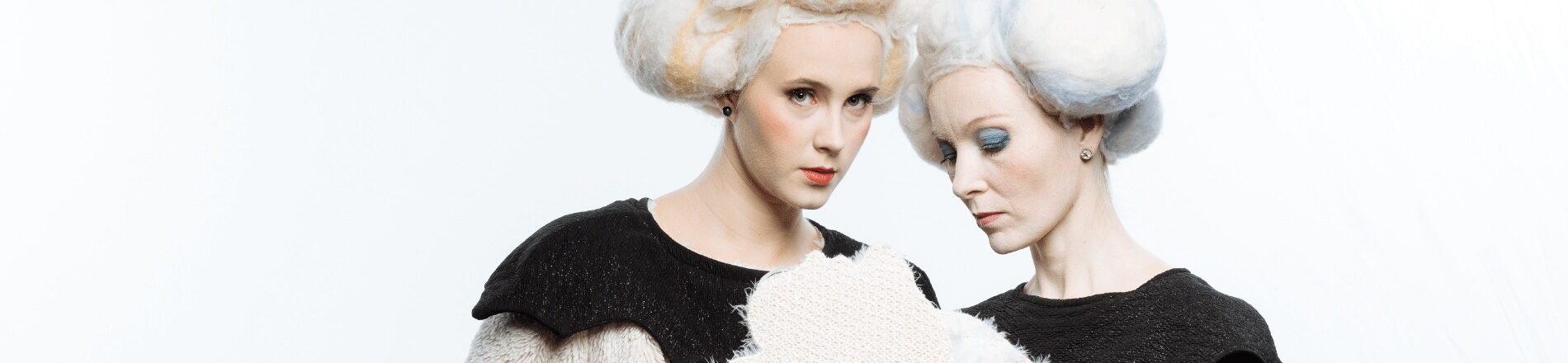Kaks naisnäitlejat suurte valgete parukatega, hoidmas käes pehmet valget nukku