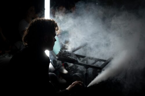 Värviline foto, mille esiplaanil on meesnäitleja, kelle peale pole valgust suunatud ning näha on vaid tema pea ja ülakeha kontuurid. Valgustatud on foto tagumine osa, kus asub lavaline konstruktsioon ning selle ümbruses uhutud lavasuits.