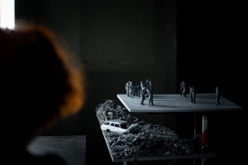 Foto laua peale asetatud miniatuursetest inimfiguuridest, mille abil on loodud stseen, mis kujutab sõja tagajärge. Halli värvi inimkujud on asetatud platoole, mille ääre all on näha ka halle (3D-prinditud) rususid. Foto vasakus ääres on aimata naisnäitleja punasejuukselist pead.