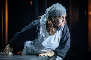 Värviline lähivaates foto naisnäitlejast, kes kannab põlle ja pearätikut ning puhastab lauda. Ta naaldub lauast eemale, suunaga küljele, pilk on suunatud kaugusesse.