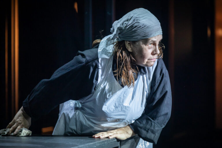 Värviline lähivaates foto naisnäitlejast, kes kannab põlle ja pearätikut ning puhastab lauda. Ta naaldub lauast eemale, suunaga küljele, pilk on suunatud kaugusesse.