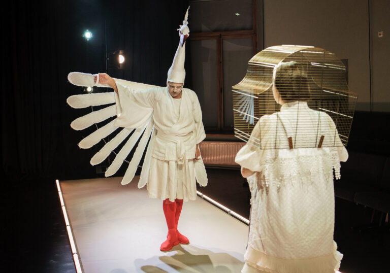 Värviline foto. Esiplaanil on seljaga kaamera poole naistegelane, kellel on seljas valge kleit ning tema pead ja õlgu ümbritseb kuldne linnupuur. Pildi keskplaanil on meestegelane, kes kannab valget kimonokleiti, punaseid sukki ja kingi ning peas on tal pika kaelaga lindu kujutav poolmask. Tal on üks käsi välja sirutatud ning selle juurest ulatub välja kostüümi osa, mis meenutab linnu tiiba. 