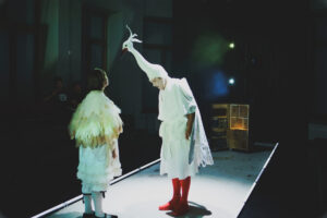 Värviline otsevaates foto, millel on üks nais- ja üks meestegelane. Naisnäitleja seisab seljaga kaamera poole, tal on seljas valge kostüüm, mille küljes on valged ja beežid suled. Meesnäitleja on valges linnukostüümis. Tal on peas pika kaelaga linnu poolmask, seljas valge kimono ning jalas punased sukad ja kingad. Mees kummardab naise poole ning naine vaatab linnu poole mehe peas. Tagaplaanil on näha avatud kohvrit, mille sees on sahtlid ja taskud. 