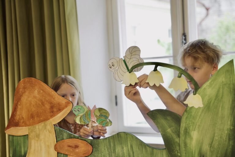 Fotol on tüdruk (vasakul) ja poiss (paremal). Nad on sirmi taga. Sirm on roheline ja selle küljes on dekoratiivsed seebed ning maikellukesed. Mõlemal lapsel on käes nukud.